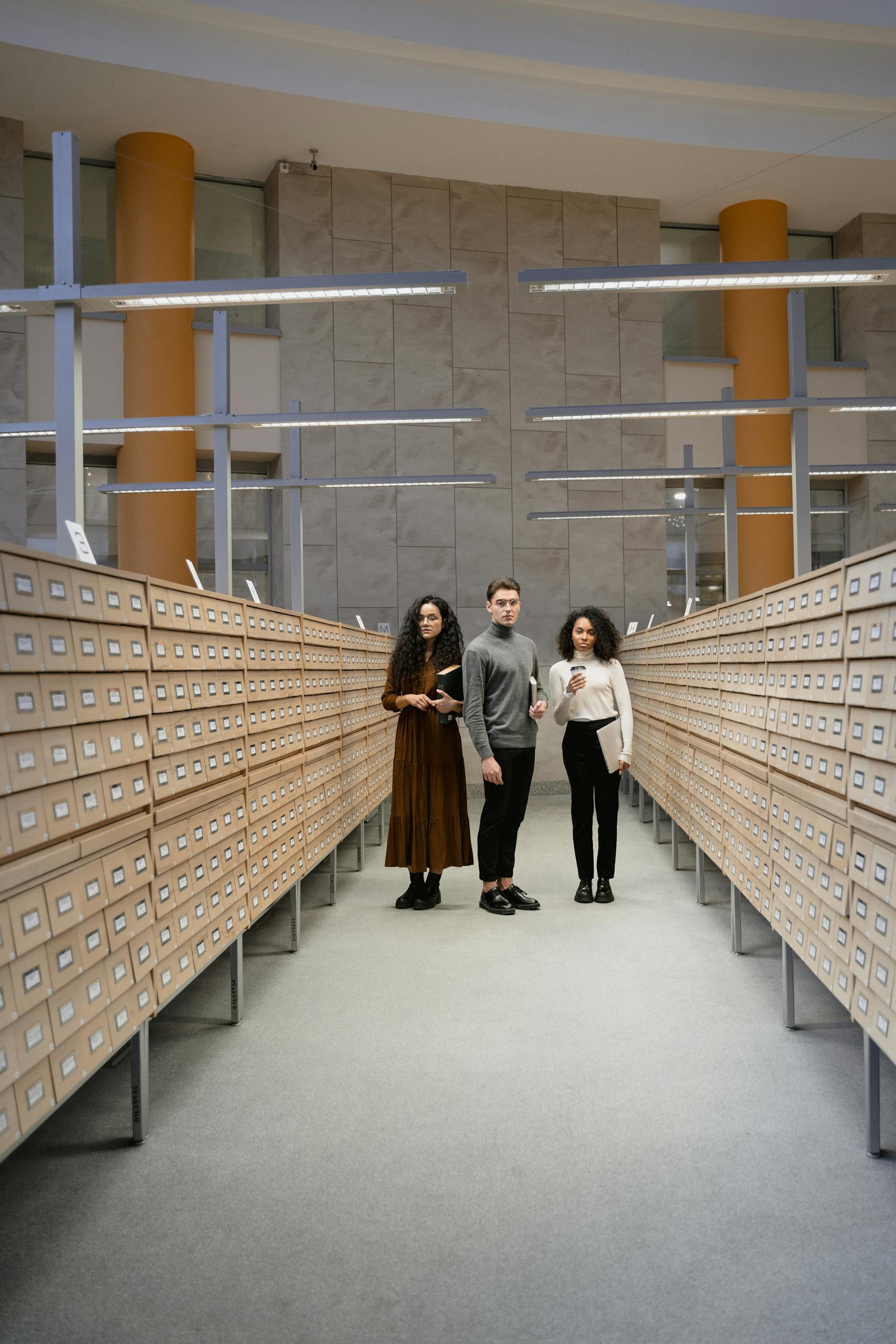 Three students standing in a library card catalog room, exploring resources for research.