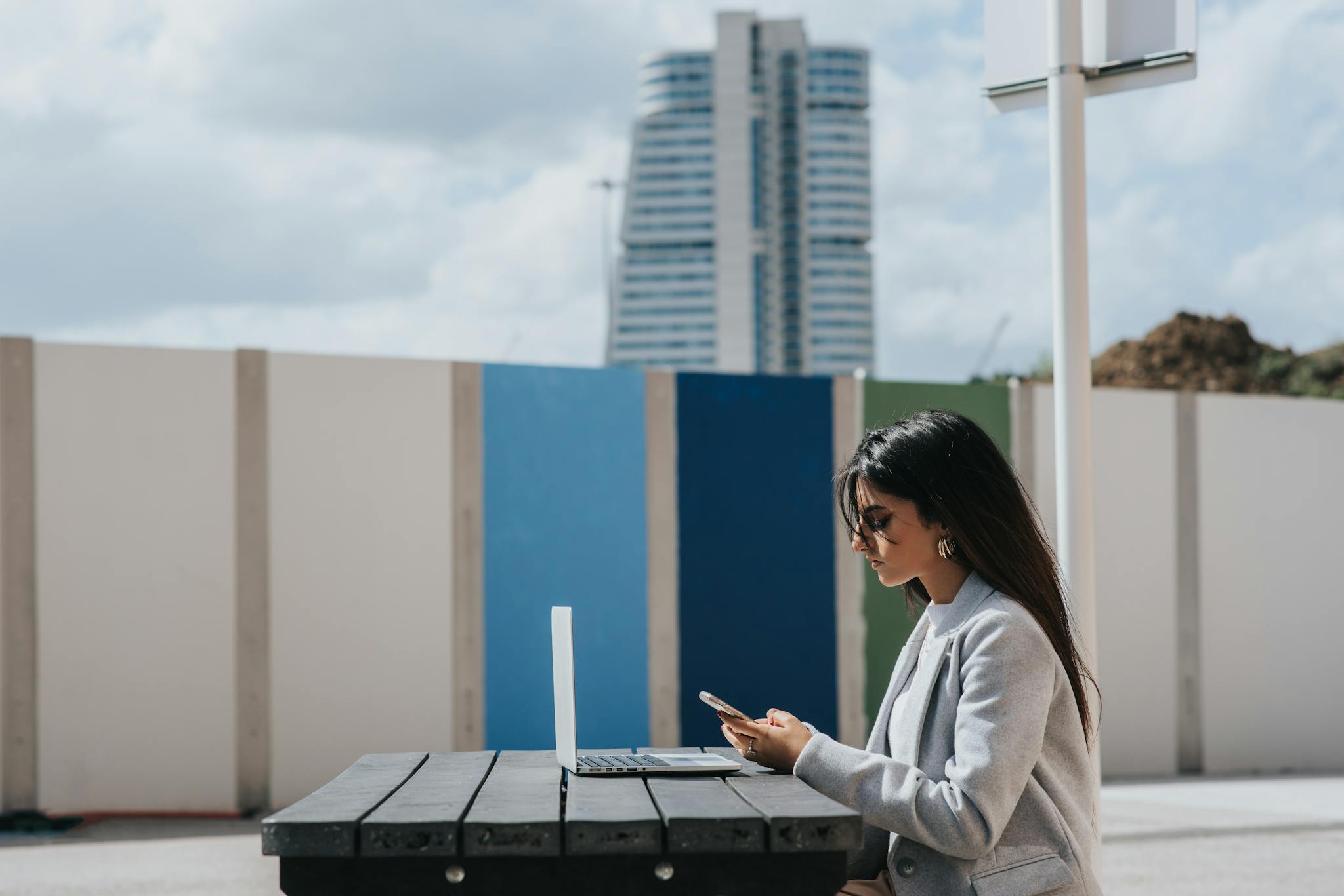 Young woman using laptop and smartphone at outdoor table. Modern cityscape background.